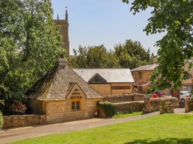 A stone building with a sloped roof near a shop and cafe sign with trees and a church tower in the background at The Bath House in Blockley