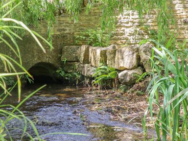 A creek flowing beside a stone wall with plants growing near an archway at The Bath House in Blockley