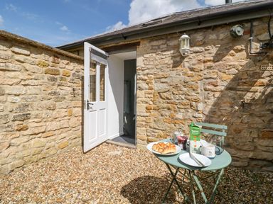 An outdoor area with a stone wall a white door open a small green table with breakfast items at The Bath House in Blockley