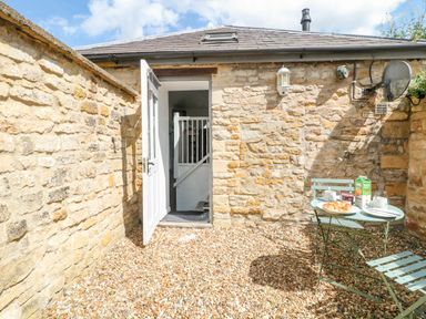 An outdoor gravel patio area with stone walls a small table and two chairs and an open door leading inside at The Bath House in Blockley