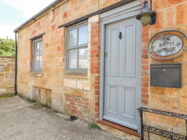 The exterior of a stone building with two windows a grey door a wall light and a sign reading The Bath House in Blockley