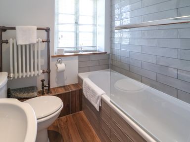 A bathroom with a bathtub, toilet, towel radiator, window with blinds and wooden floor at Red Fawn Cottage in Blockley