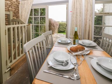 A dining table set with plates bowls cutlery glasses bread and a bottle of wine near glass doors leading to a garden at Red Fawn Cottage in Blockley
