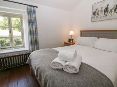 A bedroom with a bed covered with a gray blanket and white towels on top next to a window with plaid curtains at Red Fawn Cottage in Blockley