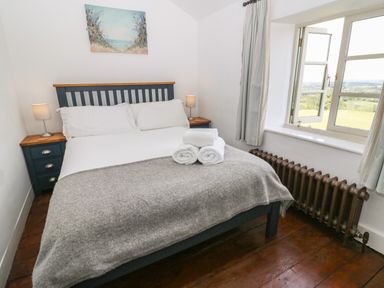 A bedroom with a double bed with white linens and gray blanket bedside tables with lamps and an open window with curtains at Red Fawn Cottage in Blockley