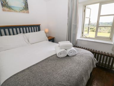 A bedroom with a bed covered in white and gray linens next to a window with an open curtain at Red Fawn Cottage in Blockley