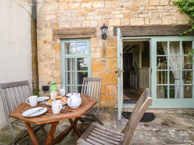 An outdoor patio with a wooden table set with cups plates croissants and a juice carton in front of a stone building with green window and door at Red Fawn Cottage in Blockley