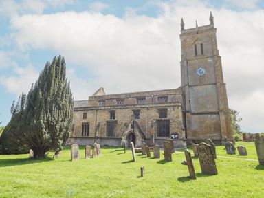 A church with a clock tower and gravestones on the grass surrounding it at Red Fawn Cottage in Blockley