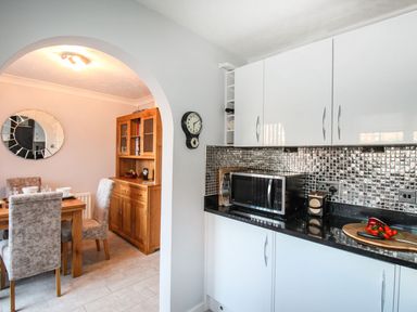 A kitchen with white cabinets a microwave and a cutting board with vegetables next to a dining area with a wooden table chairs and a wooden cabinet at Beach View House in Wyke Regis