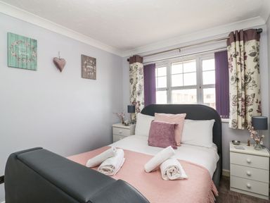 A bedroom with a bed with pink and white bedding beside two white nightstands with lamps and floral curtains at Beach View House Wyke Regis