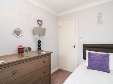 A bedroom with a wooden chest of drawers a table lamp flowers and a bed with a purple cushion at Beach View House in Wyke Regis