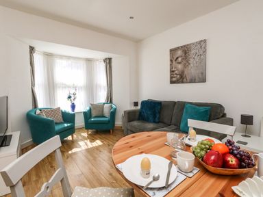 A living room with two teal armchairs, a grey sofa with pillows, a round wooden dining table set with plates and fruit bowl at Marina Reach in Weymouth