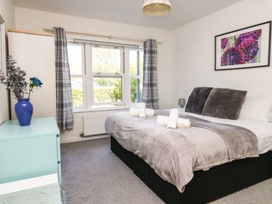 A bedroom with a double bed rolled towels a blue vase with flowers on a dresser and a window with plaid curtains at Marina Reach in Weymouth