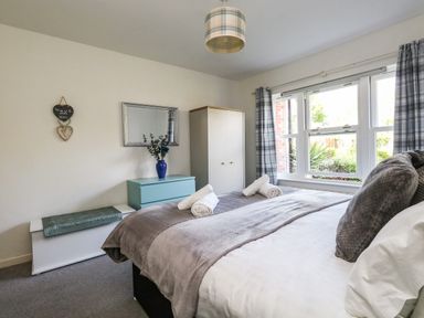 A bedroom with a bed, rolled towels, a window with curtains, a chest of drawers, a wardrobe, mirror, and a vase with flowers at Marina Reach in Weymouth