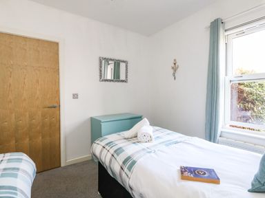 A bedroom with a bed covered in a white and plaid blanket a teal chest of drawers a wooden door a mirror and a window with teal curtains at Marina Reach in Weymouth
