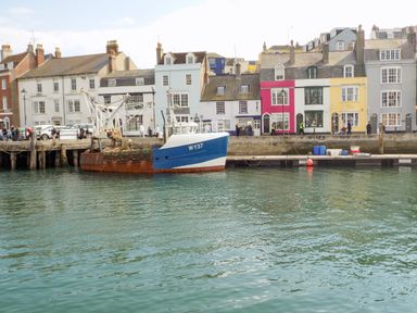 A dock with a blue and white fishing boat tied up and colorful buildings along the waterfront at Marina Reach in Weymouth