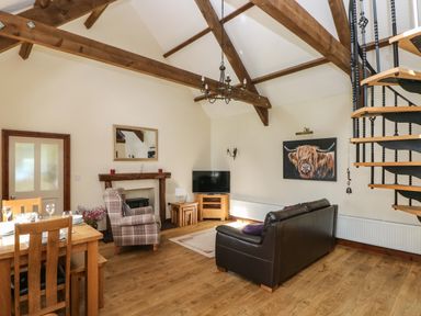 A living room with a wooden dining table chairs a plaid armchair a brown leather sofa a television a fireplace wooden beams and a spiral staircase at The Old Dairy in Maenclochog