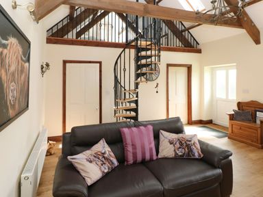 A living room with a brown leather sofa with animal print cushions a spiral staircase and wooden beams at The Old Dairy in Maenclochog