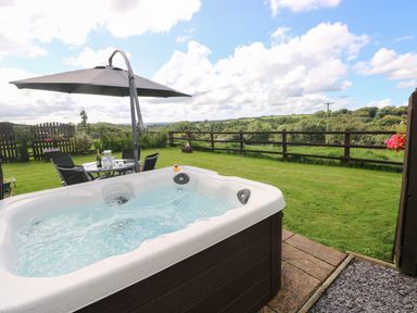 A hot tub in a garden with a table and chairs under an umbrella and a wooden fence with a view of trees at The Old Dairy in Maenclochog
