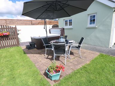 An outdoor patio area with a round glass table four chairs a large umbrella a hot tub and a flower pot on a paved surface with grass around at The Old Dairy in Maenclochog