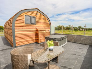 An outdoor patio with a wooden cabin hot tub wicker chairs and a glass table with flowers at Caban Cariad in Bryngwran near Bodedern