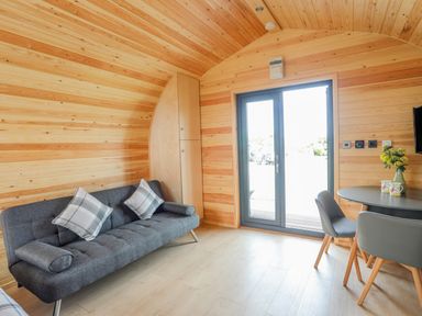 A wooden room with a gray sofa with pillows a round table with chairs and a door at Caban Cariad in Bryngwran near Bodedern