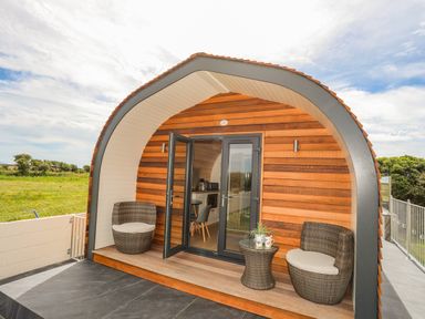An outdoor seating area with two wicker chairs and a small table in front of a wooden cabin with glass doors at Caban Cariad in Bryngwran near Bodedern