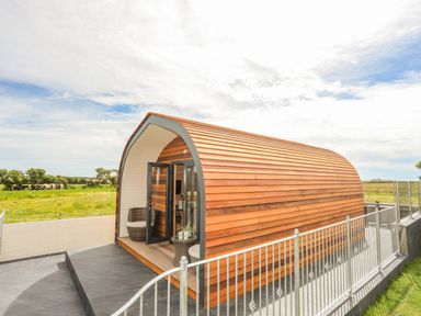 An exterior view of a wooden pod with curved roof and open glass doors showing chairs and a small table at Caban Cariad in Bryngwran near Bodedern