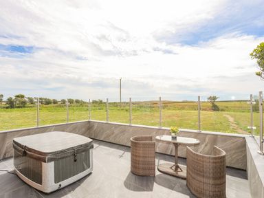 A patio with a hot tub a small glass table with two wicker chairs and grass fields beyond at Caban Cariad in Bryngwran near Bodedern