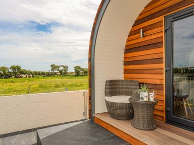 A wooden porch with a wicker chair and table with mugs and a plant overlooking a field at Caban Cariad in Bryngwran near Bodedern