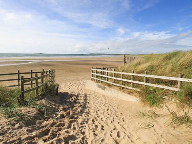 A sandy beach pathway with wooden fences leading to a wide open sandy beach and sea with blue sky at Caban Cariad in Bryngwran near Bodedern