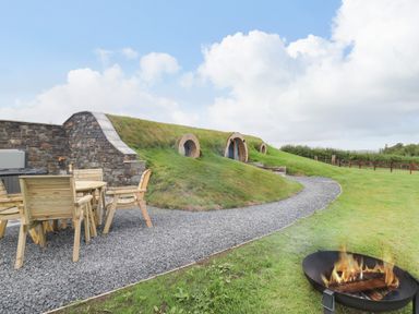An outdoor area with a fire pit wooden chairs and table on a gravel path leading to a hillside house with round windows at Tan Y Wawr - Derwen in Llanfair Caereinion