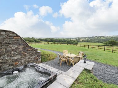 An outdoor patio with a hot tub and wooden table and chairs on gravel with a fenced grassy field in the background at Tan Y Wawr - Derwen in Llanfair Caereinion