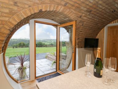 A room with a round brick ceiling and a glass door open to a stone patio with green fields beyond and a countertop with a bottle and four wine glasses at Tan Y Wawr - Derwen in Llanfair Caereinion