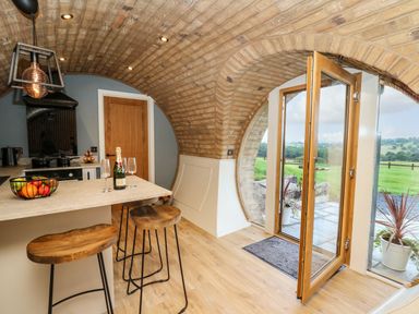 A kitchen with a curved brick ceiling wooden stools a countertop with a fruit basket and champagne bottle and an open door leading outside at Tan Y Wawr - Derwen in Llanfair Caereinion
