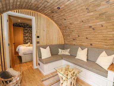A living area with a built-in corner sofa with cushions and a glass-top wooden coffee table next to a basket of blankets at Tan Y Wawr - Derwen in Llanfair Caereinion