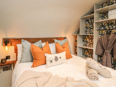 A bedroom with a bed with orange and gray pillows a wooden headboard a nightstand with an alarm clock mounted light fixtures open shelving with floral wallpaper and a hanging robe at Tan Y Wawr - Derwen in Llanfair Caereinion