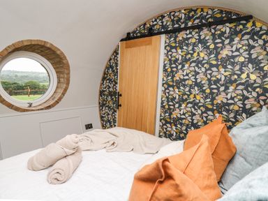 A bedroom with a round window, floral wallpaper, a wooden sliding door, and a bed with beige towels and orange pillows at Tan Y Wawr - Derwen in Llanfair Caereinion