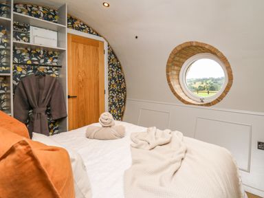 A bedroom with a bed towels and robe by a wooden door and a round window showing countryside at Tan Y Wawr - Derwen in Llanfair Caereinion