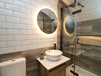 A bathroom with a round backlit mirror above a white vessel sink a toilet and a glass shower enclosure at Tan Y Wawr - Derwen in Llanfair Caereinion
