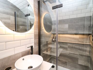 A bathroom with a round mirror a white sink a black faucet and a glass shower enclosure at Tan Y Wawr - Derwen in Llanfair Caereinion