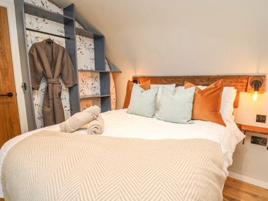 A bedroom with a bed with multiple pillows and a blanket two rolled towels on the bed a hanging robe and shelving unit near a wooden door at Tan Y Wawr - Derwen in Llanfair Caereinion
