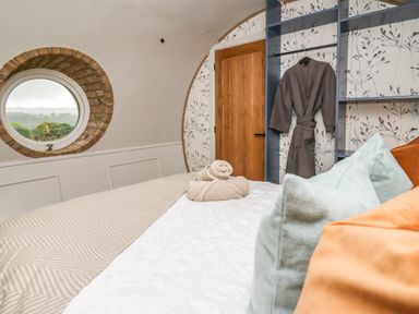 A bedroom with a round window showing a view of trees a bed with pillows and rolled towels a wooden door and a hanging bathrobe at Tan Y Wawr - Derwen in Llanfair Caereinion