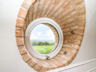 A circular window with a brick surround showing a green field and trees outside at Tan Y Wawr - Derwen in Llanfair Caereinion
