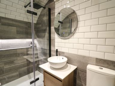 A bathroom with a shower area, round mirror, sink on a wooden cabinet, and toilet at Tan Y Wawr - Derwen Llanfair Caereinion