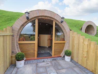 A circular wooden door set into a grass-covered hill with two potted plants outside and a wooden fence at Tan Y Wawr - Derwen in Llanfair Caereinion