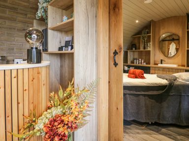A bedroom with a bed covered with a gray blanket and red and gray towels a wooden shelf with a sink and a round mirror and a wooden wall with shelves and a black handle on the door at Tan Y Wawr - Derwen in Llanfair Caereinion