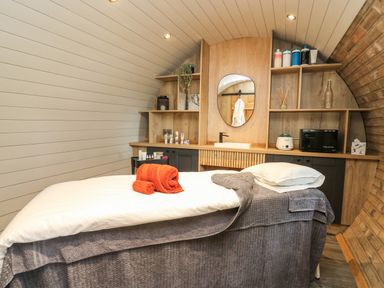 A treatment room with a massage bed covered with towels and a pillow and wooden shelves with beauty products at Tan Y Wawr - Derwen in Llanfair Caereinion