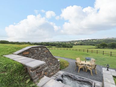 An outdoor patio area with a hot tub wooden table and chairs overlooking a grassy field and hills at Tan Y Wawr - Derwen in Llanfair Caereinion