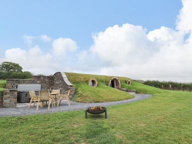 An outdoor area with a wooden table and chairs next to a stone wall and hot tub and a house built into a grassy hill at Tan Y Wawr - Derwen in Llanfair Caereinion